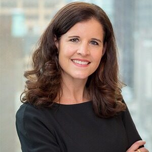 A portrait of Katie Donald. She is a white woman wearing a wide-neck black blouse and has long, loosely curled brown hair.