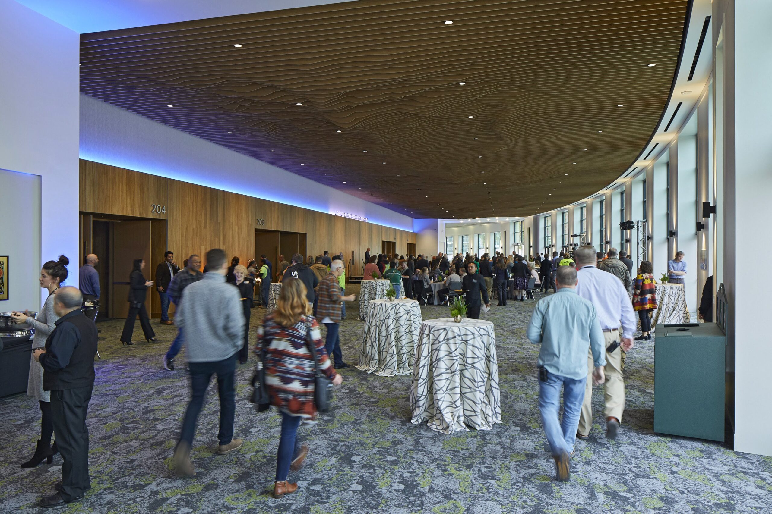 The pre-function ballroom at the Oregon Convention Center. Attendees are walking through the space in between small tables. Blue lighting runs along the wood paneled ceiling.