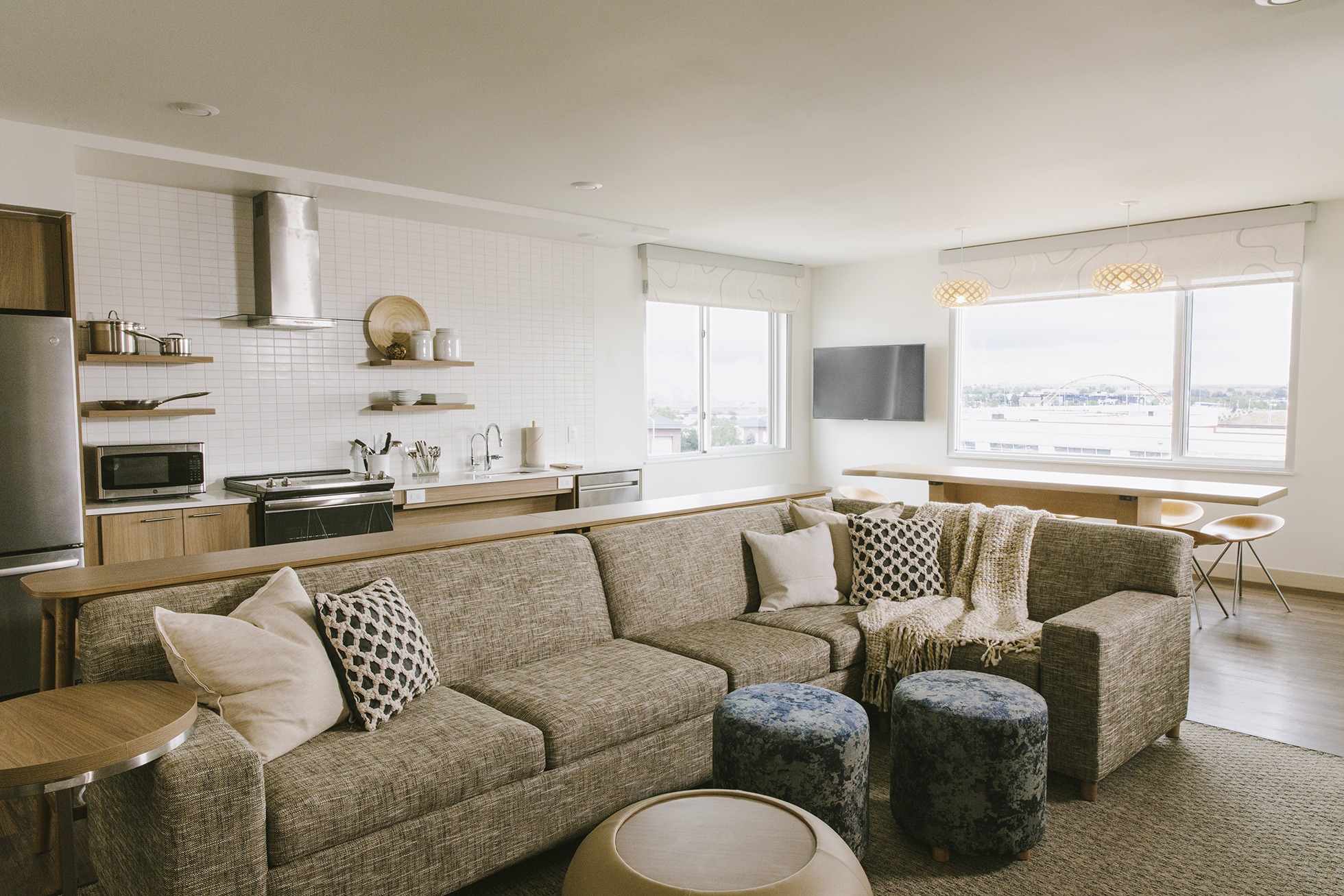 A living room in an Element by Westin hotel. A tan couch with earth-tone pillows is in front of a kitchen with white subway tiles.