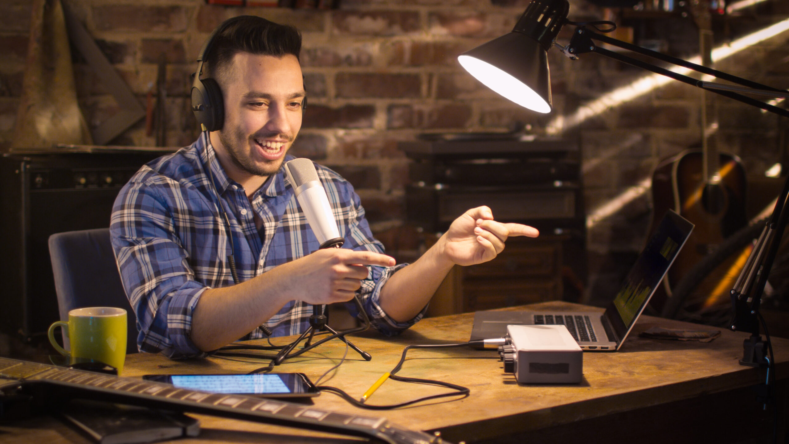 A man sits at a desk with a laptop, microphone, and headphones. 
