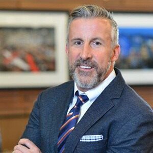 A portrait of Ron Bracco. He is an older white man with combed grey hair, a short beard and a grey suit.