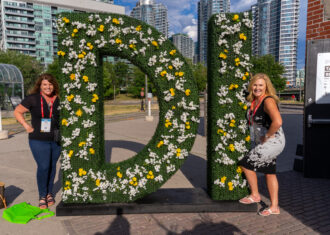 two women posing next to a floral sign that says "DI"