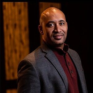 A portrait of Derek Douglass Drew. He is a bald black man with a grey suit and a red collared shirt.