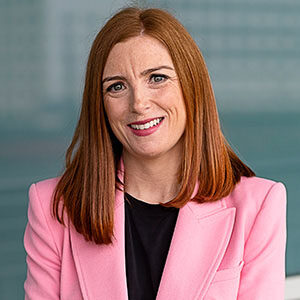 A portrait of Michelle Kemp. She is a white woman with long red hair and a pink suit jacket