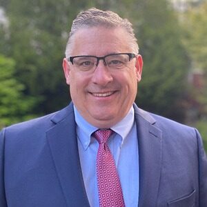 A portrait of Mark Goeman. He is a white man with square glasses, a grey suit and a red tie.