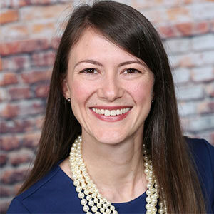 A portrait of Megan Martin. She is a white woman with long dark hair, a pearl necklace and a dark blue blouse