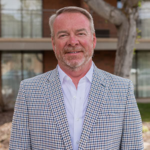 A portrait of Anthony Moody. He is an older white man with combed hair, a stubbled beard and a patterned suit 