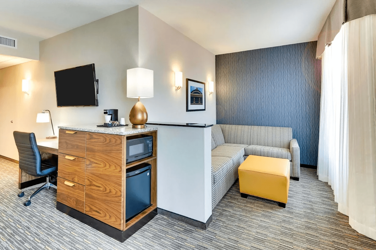 The interior of a suite at Drury Plaza Hotel Orlando. A small seating area is around a corner next to a window. A TV and cabinet are next to a desk.