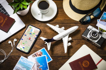 Top view of an tiny airplane surrounded by various traveling stuff such as a coffee cup, a camera with some snapshots, a smartphone with a map on the screen, an opened notebook, two passports with boarding passes and some summer accessories like a hat and sunglasses. All the objects are on a rustic wooden desk