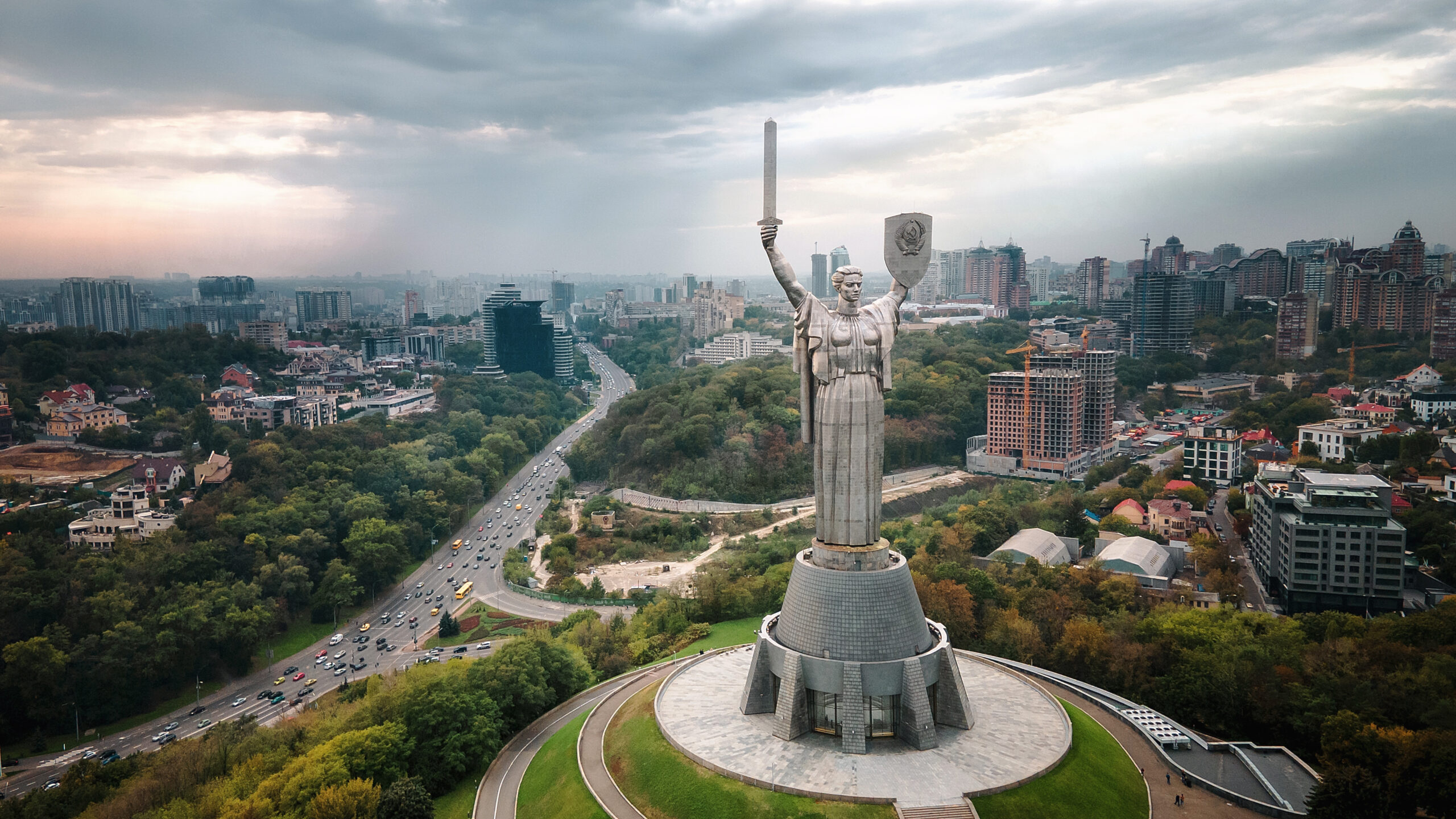 The Motherland Monument in Kyiv, a statue of a woman holding a sword and shield up. The city skyline is behind her.