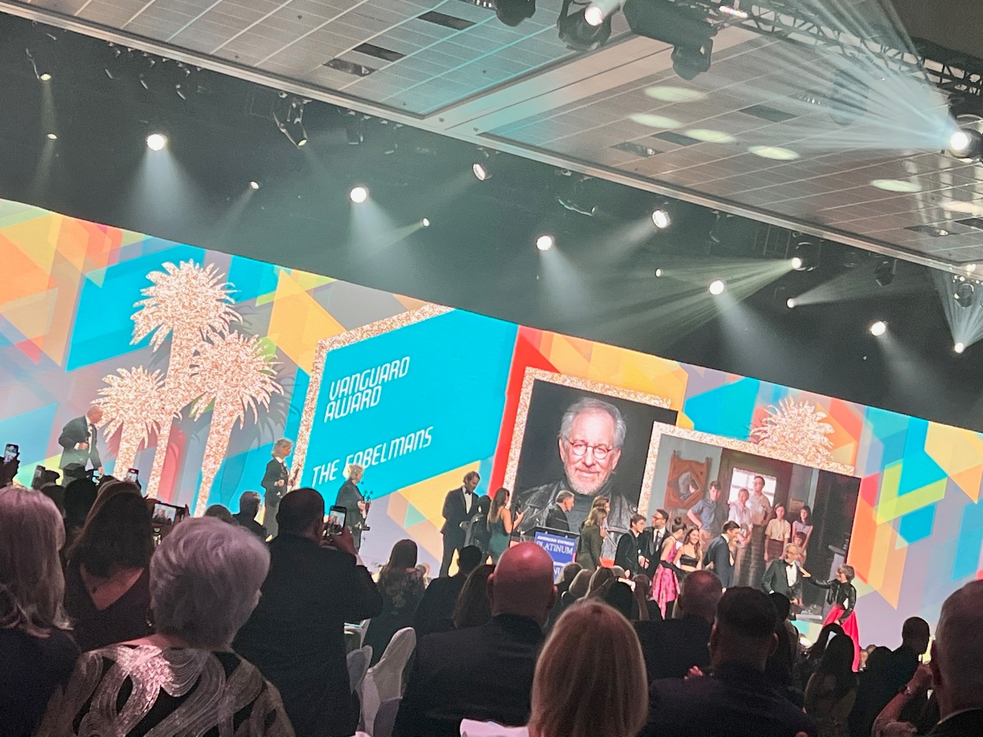 cast on stage at the vanguard awards in palm springs, california. On screen is an image of steven speilberg and a still shot from his movie, "The Fabelmans"