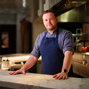 Chef Michael Yates standing with palms on table, wearing dark blue apron and light blue dress shirt
