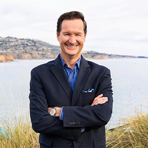 troy wood with arms crossed standing in front of body of water, wearing black sport coast and blue dress shirt