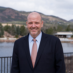 man standing in front of lake, wearing black sport coat and blue dress shirt