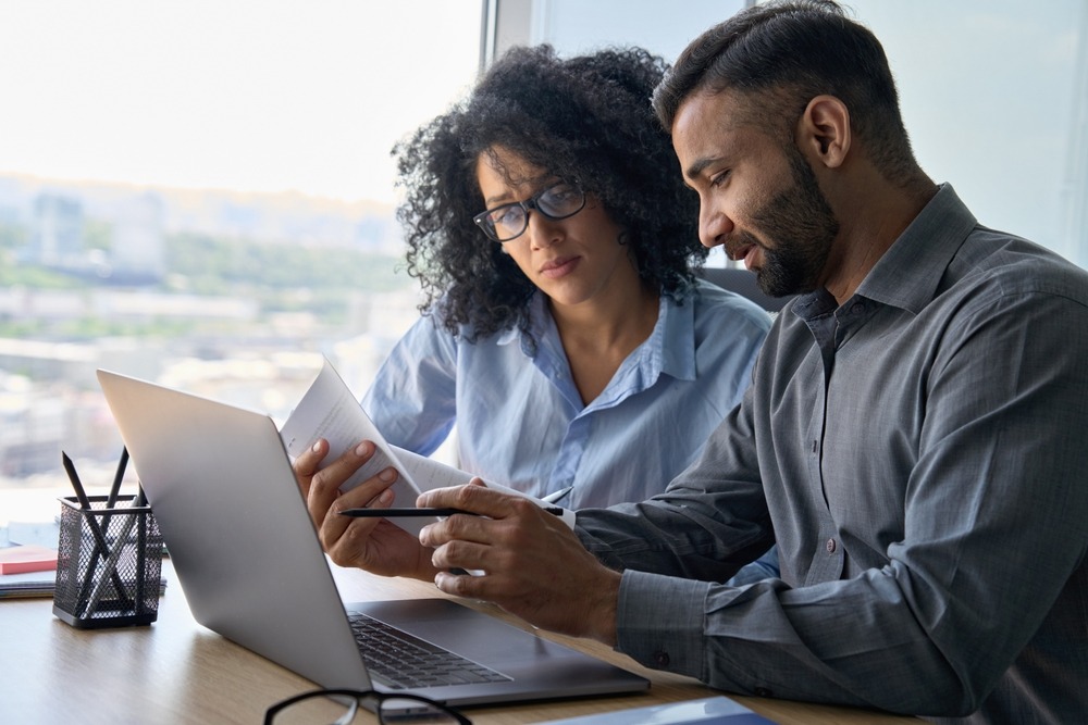 Multiethnic male indian mentor and female African American sitting at desk with laptop doing paperwork together