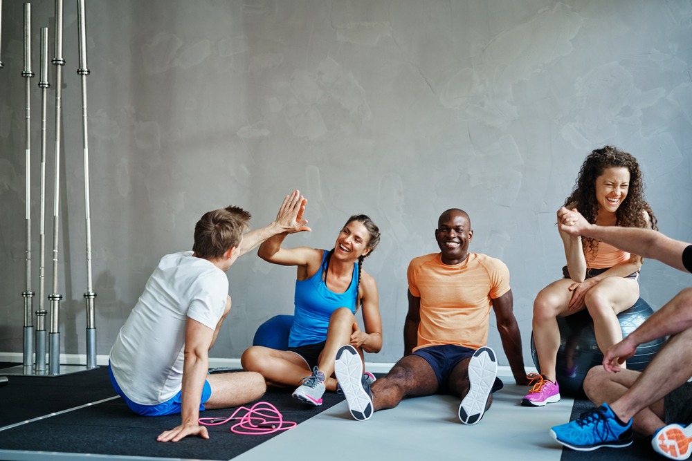 Friends in sportswear high fiving and laughing together while sitting on the floor of a gym after a workout