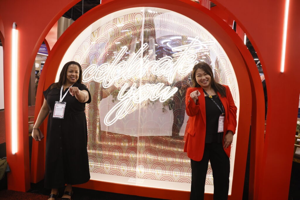 two attendees at IMEX pointing in front of Celebrate You neon