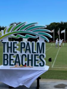 Three people in white playing croquet at National Croquet Center behind a The Palm Beaches sign