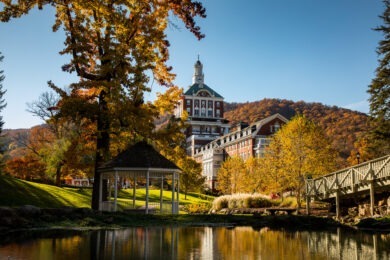 The Omni Homestead Resort stands next to a lake and trees in front of hills with fall foliage