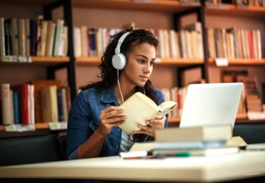 woman in library with book in her hands and headphones on looking at computer