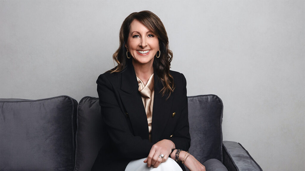 Kelly Gleeson Smith sitting on couch in black sport coat and gold silk shirt