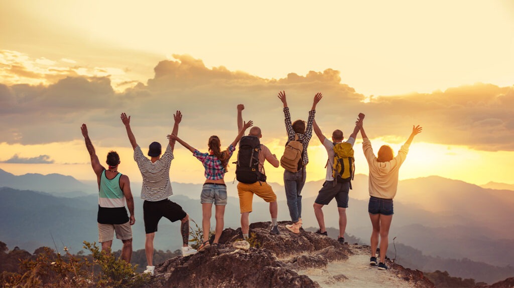 Group of happy friends standing with raised arms on mountain at sunset