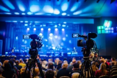 Two professional video cameras on tripods recording a concert with an audience in the foreground